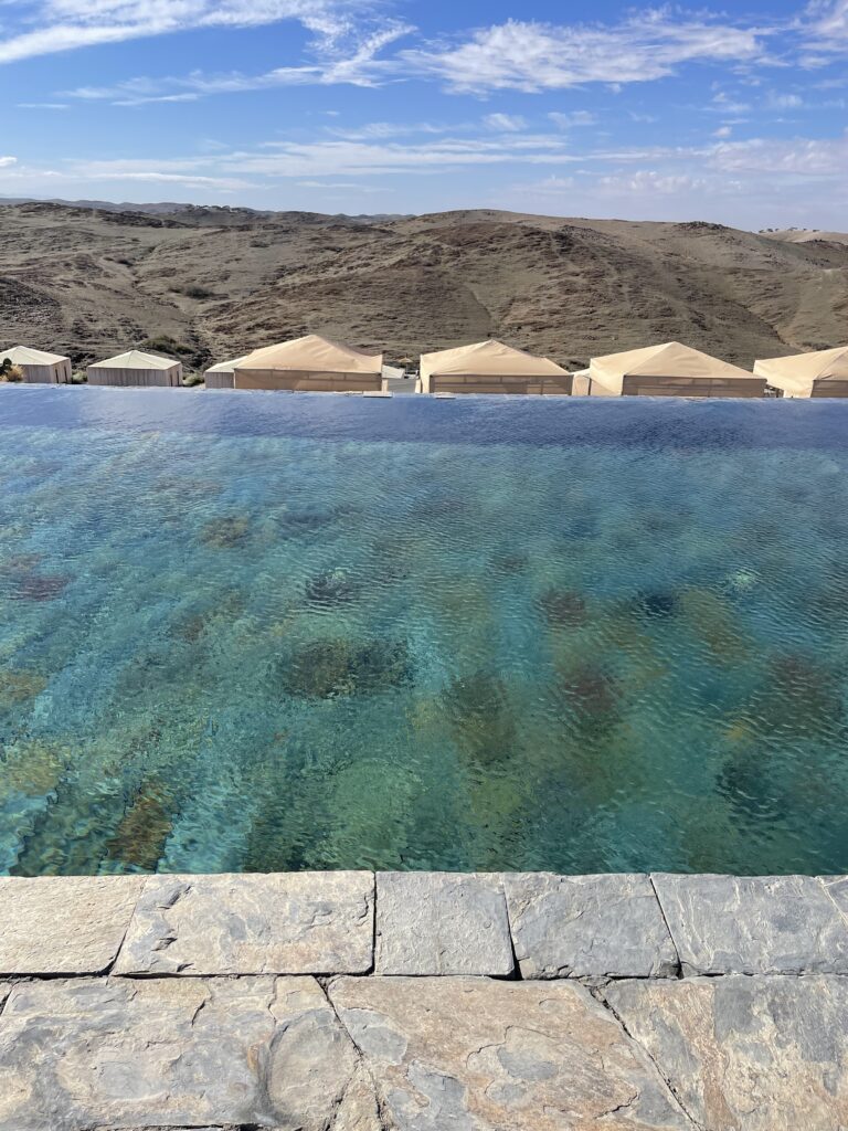 An infinity pool at The White Camel Agafay overlooking a serene desert landscape with traditional Moroccan tented lodgings in the background, under a clear blue sky.