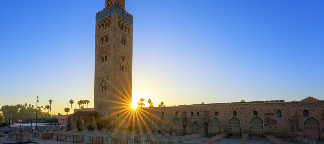 Sunrise behind the Koutoubia Mosque in Marrakech with a clear blue sky, highlighting the mosque's silhouette and palm trees.