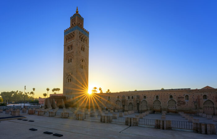 Sunrise behind the Koutoubia Mosque in Marrakech with a clear blue sky, highlighting the mosque's silhouette and palm trees.