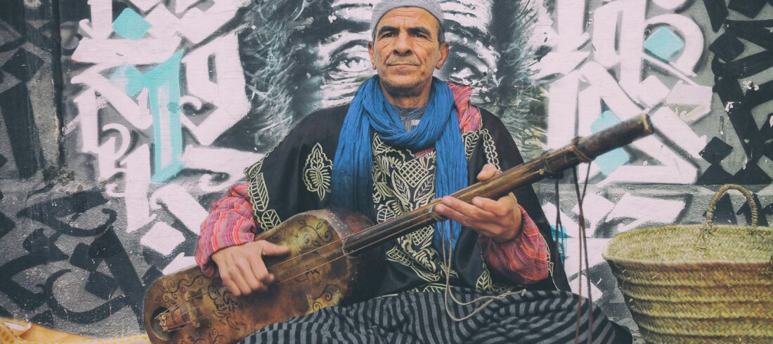 A seasoned Gnawa musician in traditional attire playing a guembri against an urban graffiti backdrop in Morocco.