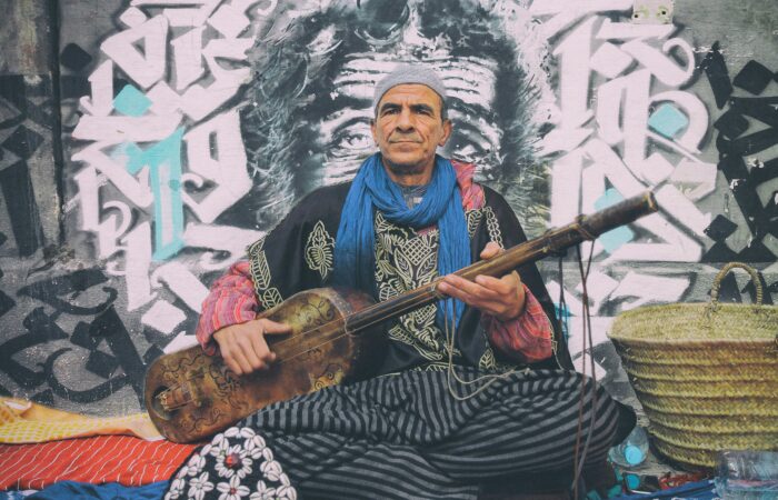 A seasoned Gnawa musician in traditional attire playing a guembri against an urban graffiti backdrop in Morocco.