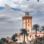 View of a traditional Moroccan tower with a lighthouse against a cloudy sky, surrounded by lush palm trees near the ocean from Maroc