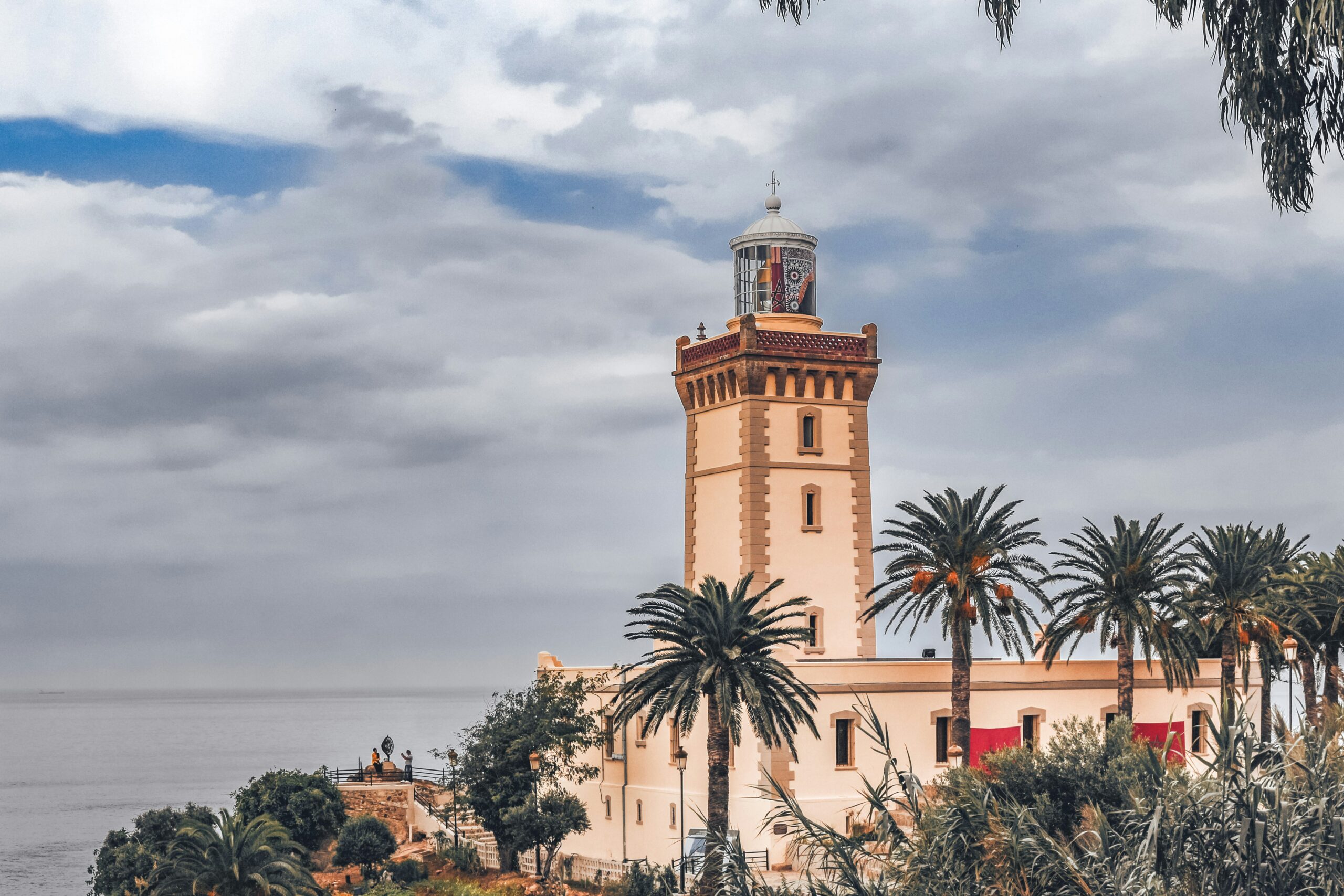 View of a traditional Moroccan tower with a lighthouse against a cloudy sky, surrounded by lush palm trees near the ocean from Morocco