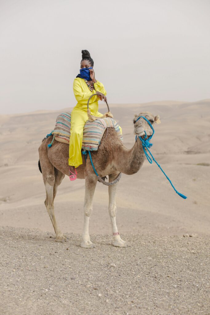 A person in vibrant yellow attire and a blue face covering sits atop a camel with a multicolored saddle in the Agafay Desert, with rolling sandy hills at the agafay desert stretching into the distance under a hazy sky.