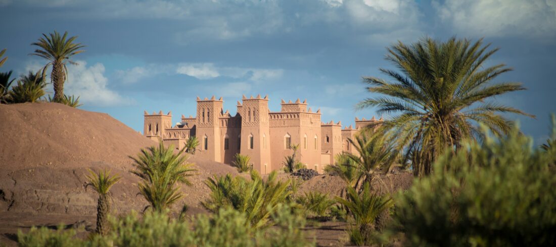 A stunning Kasbah stands with earthen walls and ornate towers against a backdrop of blue skies, lush palm trees, and the undulating terrain of the High Atlas Mountains.
