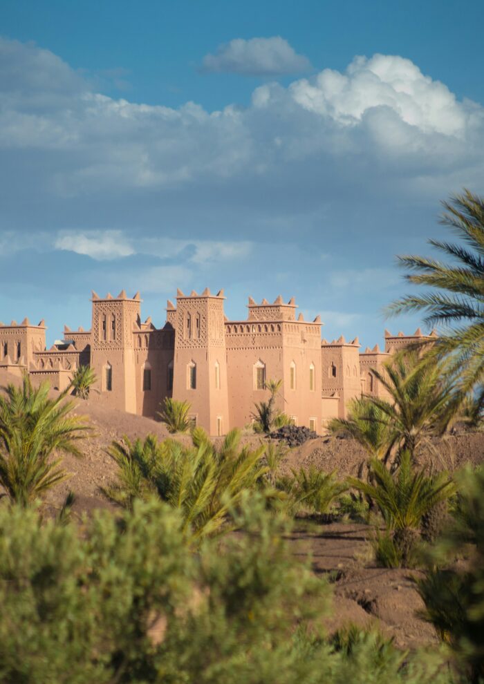 A stunning Kasbah stands with earthen walls and ornate towers against a backdrop of blue skies, lush palm trees, and the undulating terrain of the High Atlas Mountains.